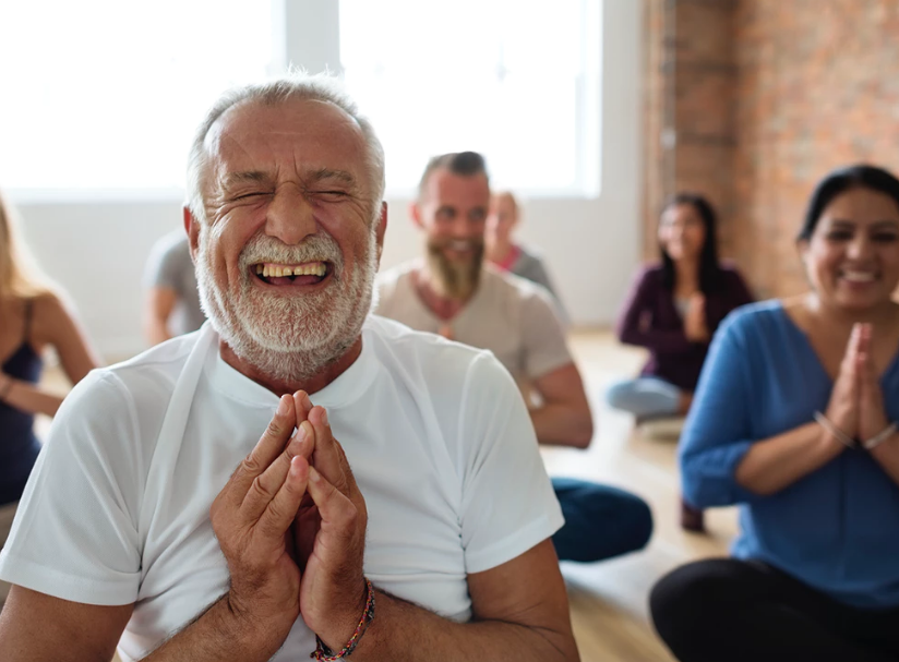 old man in white shirt smiling and squinting with eye closed doing yoga and laughing mind body and soul good for the heart stock photo