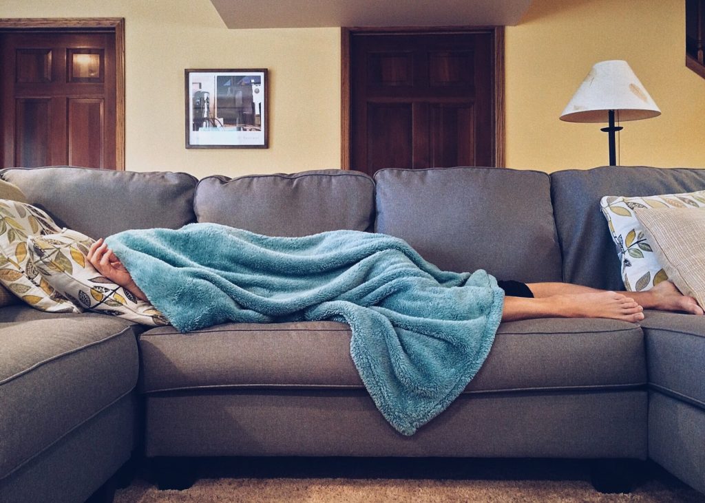 white woman hiding under blue blanket on grey gray sectional couch sick at home watching super bowl