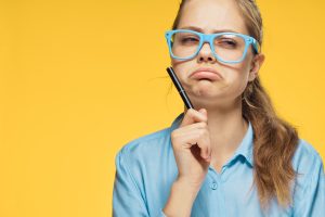 young woman frowning and looking to the side in contemplation in a blue shirt and glasses on a yellow background