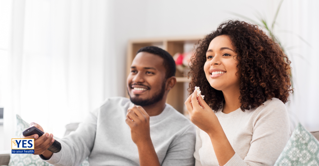 man and woman smiling, eating popcorn, and watching tv