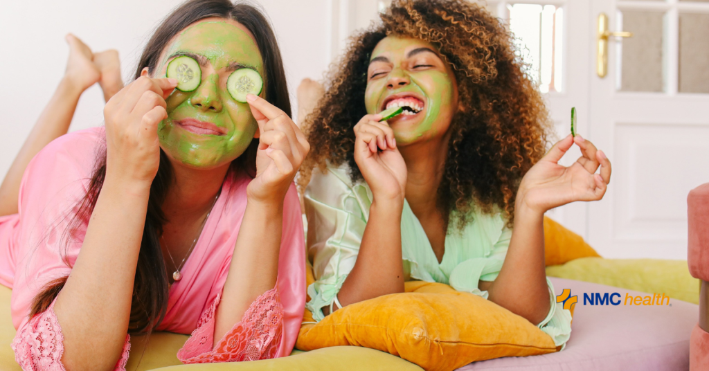 two smiling young women in robes performing skincare routine with green face masks and cucumbers