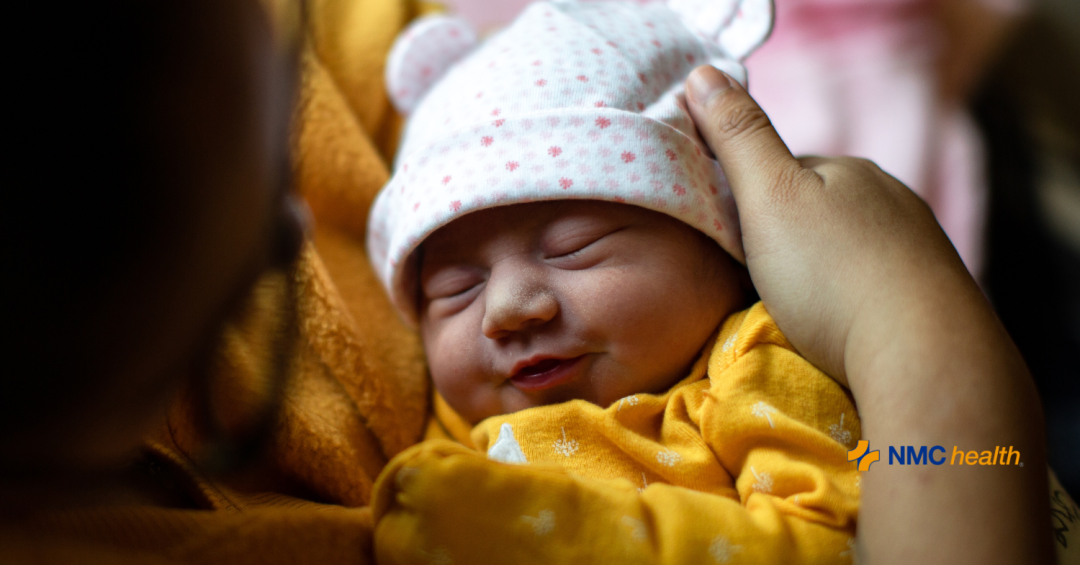 young mom holding smiling newborn wearing yellow onesie and white cap to her chest