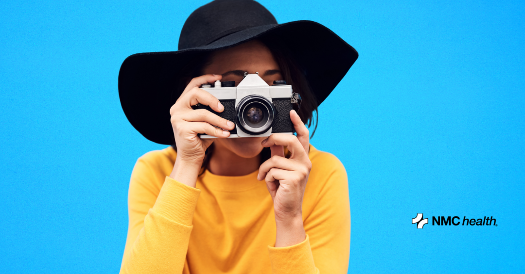 woman in yellow shirt and black hat holding camera up to her face taking a photo on blue background